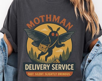 May include: A dark gray t-shirt with a vintage-style illustration of a mothman with red eyes carrying a package. The text "Mothman Delivery Service" is printed in orange and brown, with the tagline "Fast. Silent. Slightly Ominous."