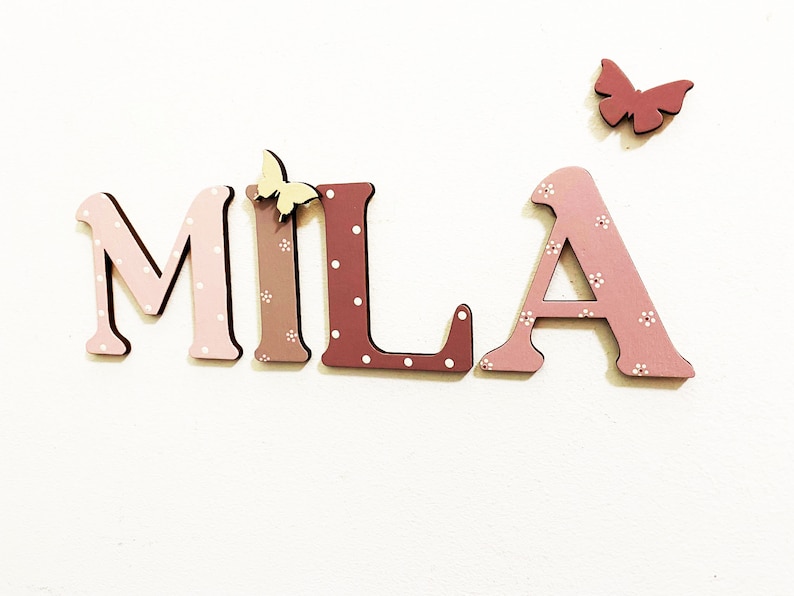 Pode incluir: Letras de madeira que soletram o nome "MILA" em rosa e marrom com bolinhas brancas. As letras s&atilde;o decoradas com pequenas borboletas amarelas.
