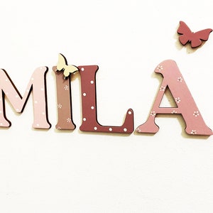 Pode incluir: Letras de madeira que soletram o nome "MILA" em rosa e marrom com bolinhas brancas. As letras s&atilde;o decoradas com pequenas borboletas amarelas.