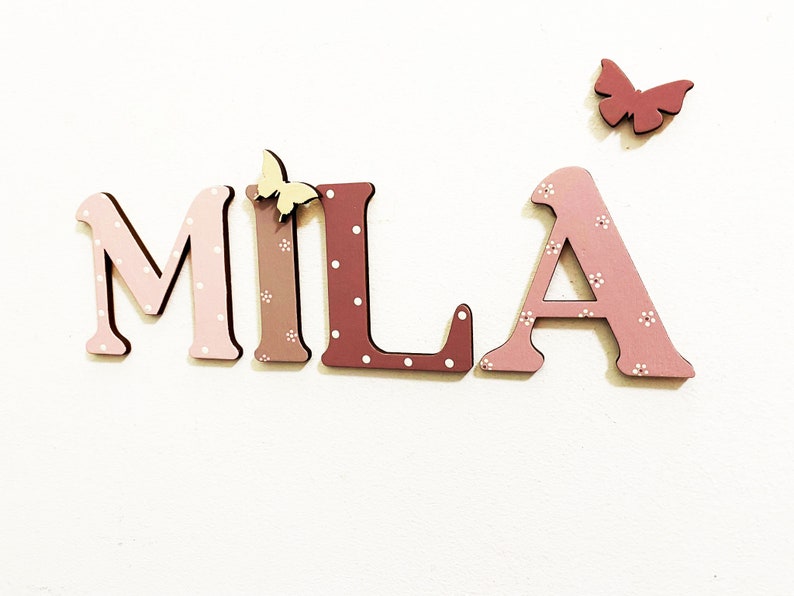 Pode incluir: Letras de madeira que soletram o nome "MILA" em rosa e marrom com bolinhas brancas. As letras s&atilde;o decoradas com pequenas borboletas amarelas.