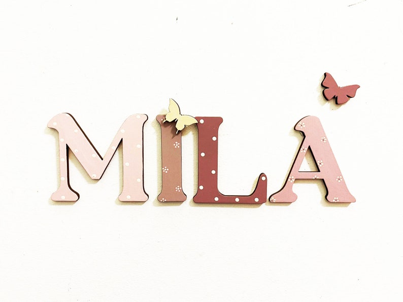 Pode incluir: Letras de madeira que soletram o nome "MILA" em rosa e marrom com bolinhas brancas. As letras s&atilde;o decoradas com pequenas borboletas amarelas e vermelhas.