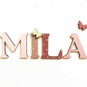 Pode incluir: Letras de madeira que soletram o nome "MILA" em rosa e marrom com bolinhas brancas. As letras s&atilde;o decoradas com pequenas borboletas amarelas e vermelhas.