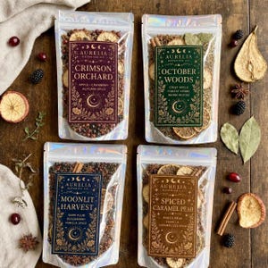 May include: Four clear stand-up pouches containing dried botanical potpourri blends labeled Aurelia Botanica Co. The labels feature celestial motifs and names: Crimson Orchard, October Woods, Moonlit Harvest, and Spiced Caramel Pear.
