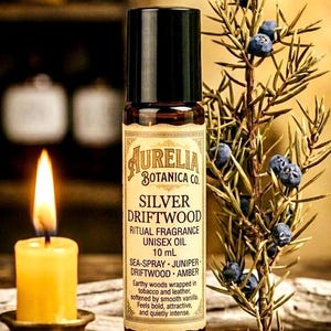 May include: Aurelia Botanica Co. Silver Driftwood unisex ritual fragrance oil in a 10 ml bottle. The label lists sea-spray, juniper, driftwood, and amber notes. A lit candle and a sprig of berries are in the background.