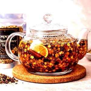 May include: A clear glass teapot filled with tea, orange slices, and cinnamon sticks sits on a round cork coaster. Beside it is a silver pouch labelled Prosperity & Abundance Tea, a small bowl of loose tea, and a pile of dry tea leaves.