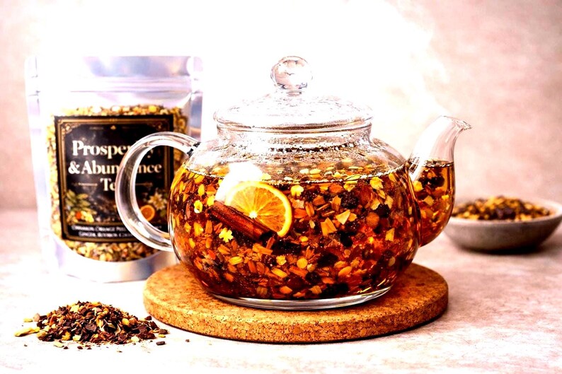 May include: A clear glass teapot filled with tea, orange slices, and cinnamon sticks sits on a round cork coaster. Beside it is a silver pouch labelled Prosperity & Abundance Tea, a small bowl of loose tea, and a pile of dry tea leaves.
