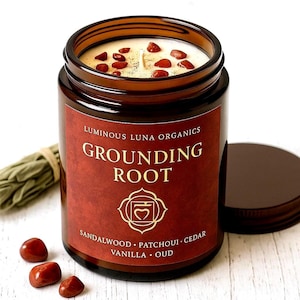 May include: A brown glass jar candle with a red label that reads "Grounding Root" and lists ingredients. The candle has a white wax top with red stones. A bundle of sage and loose stones are next to the candle.