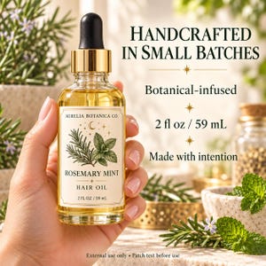 K&ouml;nnte beinhalten: Eine klare Glasflasche mit Rosemary Mint Haar&ouml;l, schwarzem Tropfverschluss und goldenem Kragen. Das Etikett zeigt botanische Illustrationen, den Text Aurelia Botanica Co., Rosemary Mint Hair Oil und 2 fl oz / 59 mL. Der Text lautet Handcrafted in small batches, Botanical-infused, 2 fl oz / 59 mL, Made with intention sowie External use only &bull; Patch test before use.