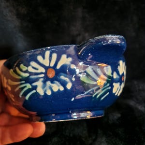 Peut inclure: Petit bol en céramique bleu avec une anse. Le bol est décoré de motifs floraux blancs et jaunes. Le bol est tenu dans une main, et le fond est sombre.