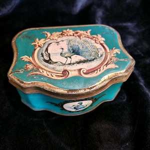 May include: A teal metal storage tin with a scalloped edge and gold-toned trim. The lid features a central oval illustration of a peacock with its tail fanned out, surrounded by ornate gold floral scrollwork on a teal background.