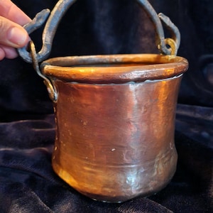 Puede incluir: Un pequeño cubo de cobre antiguo con un asa de hierro curvada. El cubo tiene una base redondeada y un borde ligeramente ensanchado. El asa está sujeta con detalles decorativos y rizados. El cobre tiene una rica pátina envejecida, dándole un aspecto vintage.