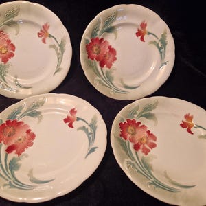 Puede incluir: Cuatro platos de cerámica blanca con un diseño floral. Cada plato presenta una flor roja y amarilla con hojas y tallos verdes. Los platos tienen un borde festoneado.