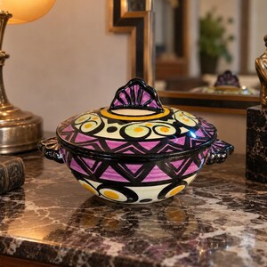 Peut inclure: Une soupière en céramique avec couvercle, ornée d'un motif géométrique peint à la main dans des tons violet, noir, jaune et crème. Le bol possède deux anses latérales et un couvercle décoratif avec un bouton central, aux lignes marquées.