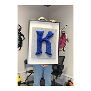 Może przedstawiać: Ramka z obrazem przedstawiającym dużą, niebieską, puszystą literę "K" na szarym tle.