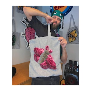 May include: A gray tote bag with a large, textured moth design in shades of pink, gray, and brown. The bag has two handles and is held up by a person. Other similar bags with insect designs are visible in the background.