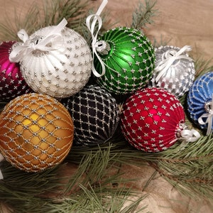 Op de afbeelding: Een verzameling van negen kerstballen met kralen in verschillende kleuren en patronen, waaronder goud, rood, groen, wit en blauw. De kerstballen zijn gerangschikt op een bed van dennennaalden.
