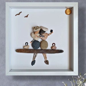 May include: A framed pebble art piece depicting a couple sitting on a driftwood bench. The couple is made of small, smooth stones in various colours and shapes. The background is white with a small gold sun and two brown birds.
