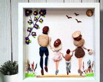 May include: A square white framed wall art piece featuring a family of four created from smooth pebbles and stones. The figures are arranged in a line against a white background, with a purple flower branch, three birds, and a moon above them.
