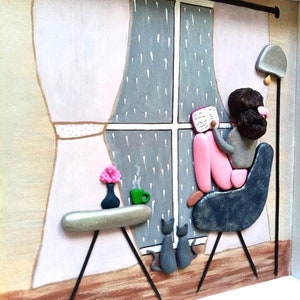 May include: A framed pebble art piece depicting a person sitting in a dark blue chair reading a book by a window with rain falling outside. A small table holds a green mug and a vase with pink flowers, while two gray cats sit on the floor.