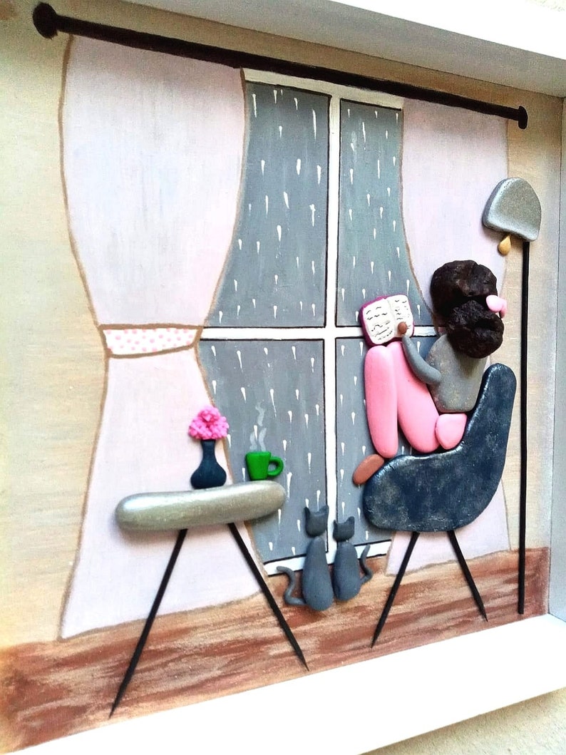May include: A framed pebble art piece depicting a person sitting in a dark blue chair reading a book by a window with rain falling outside. A small table holds a green mug and a vase with pink flowers, while two gray cats sit on the floor.