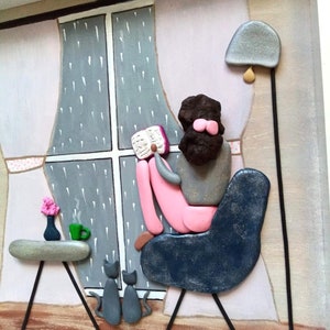 May include: A pebble art piece depicting a person sitting in a dark blue chair reading a book by a window with rain falling outside. Two gray cats sit on the floor, and a small table holds a green mug and a vase with pink flowers.