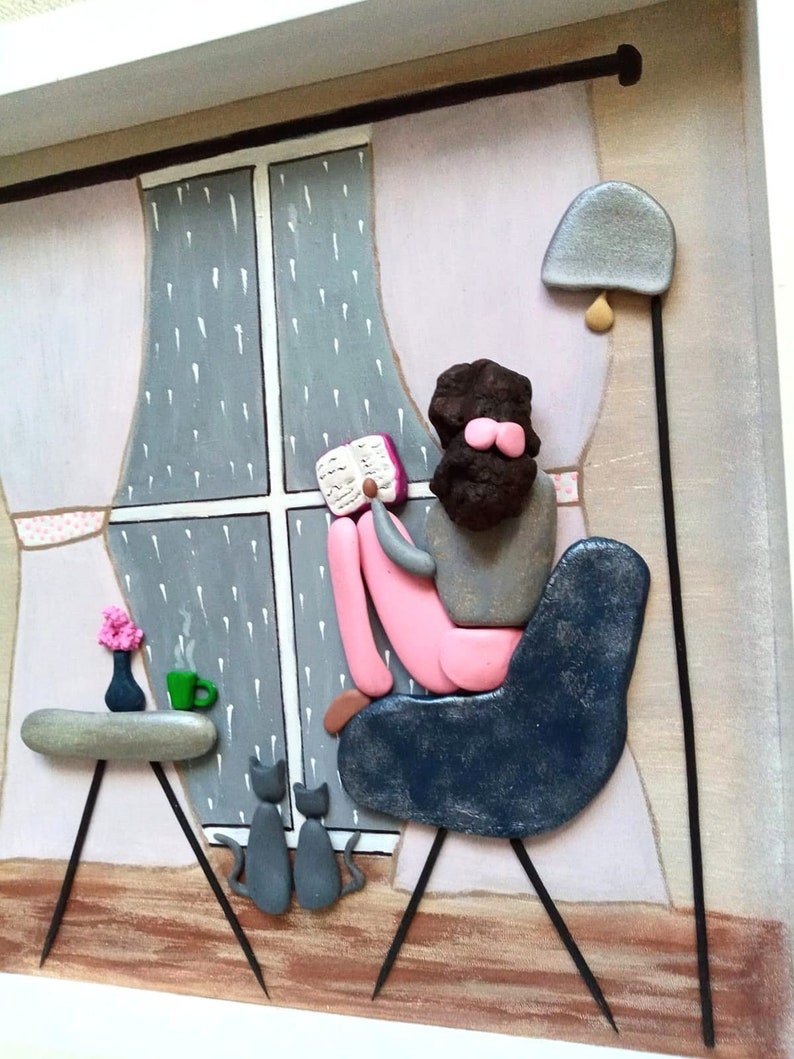 May include: A pebble art piece depicting a person sitting in a dark blue chair reading a book by a window with rain falling outside. Two gray cats sit on the floor, and a small table holds a green mug and a vase with pink flowers.