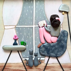 May include: A pebble art piece depicting a figure with dark hair sitting in a dark blue chair reading a book by a window with rain. Two gray cats sit on the floor. A side table holds a small vase with pink flowers and a green mug with steam.