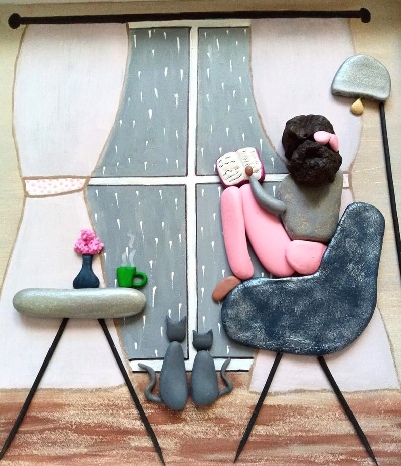 May include: A pebble art piece depicting a figure with dark hair sitting in a dark blue chair reading a book by a window with rain. Two gray cats sit on the floor. A side table holds a small vase with pink flowers and a green mug with steam.