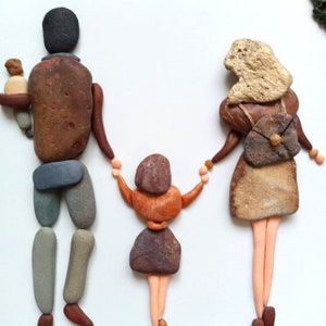 May include: A pebble art piece depicting a family of three figures. The left figure has a dark head and brown torso, holding a small baby. The center figure is a child with a brown head and orange torso. The right figure has a light head and brown dress.