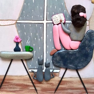 May include: A pebble art piece depicting a figure with dark hair sitting in a dark blue chair reading a book by a window with rain. Two gray cats sit on the floor. A small table holds a green mug and a vase with pink flowers. The scene is set against a painted background with light pink curtains.