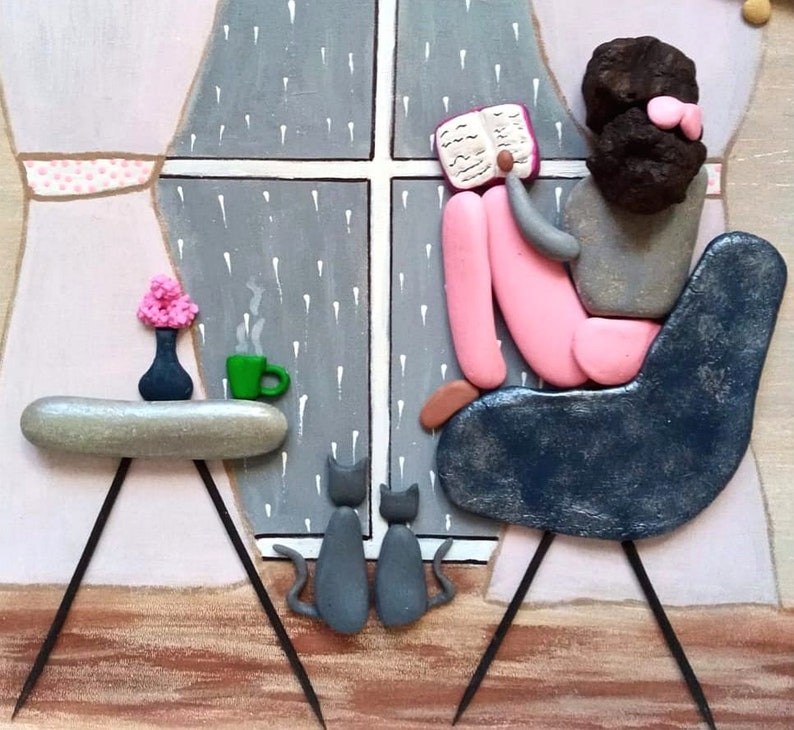 May include: A pebble art piece depicting a figure with dark hair sitting in a dark blue chair reading a book by a window with rain. Two gray cats sit on the floor. A small table holds a green mug and a vase with pink flowers. The scene is set against a painted background with light pink curtains.