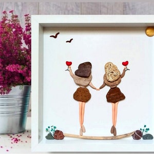 Könnte beinhalten: Ein gerahmtes Kunstwerk mit zwei Figuren aus Steinen, die rote Herzen halten und auf einem Treibholzast stehen. Das Kunstwerk ist vor einem weißen Hintergrund mit einer kleinen Sonne oben rechts platziert.
