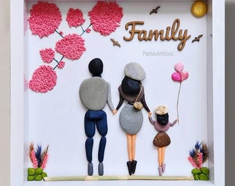 May include: A framed pebble art piece depicting a family of four. The artwork features pebble figures holding hands, with a wooden "Family" sign and pink floral accents. The background is white, and the frame is square.