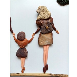 May include: A pebble art piece depicting two figures holding hands, crafted from smooth brown and tan stones. The larger figure has a textured gold-toned stone head and a backpack, while the smaller figure has a dark brown stone head.