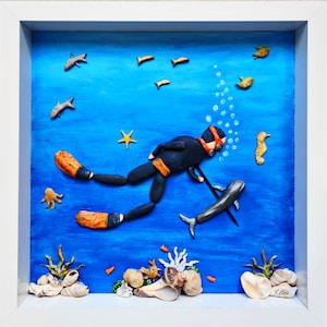 May include: A framed artwork depicting a scuba diver made of stones, swimming in a blue ocean with other sea creatures. The diver is holding a spear and is surrounded by fish, a starfish, a seahorse, and bubbles. The artwork is framed in white and has a blue background.