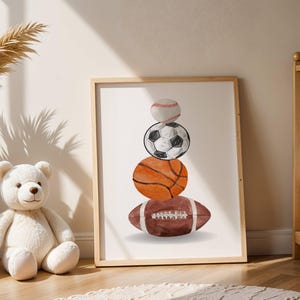 Pode incluir: Uma impress&atilde;o emoldurada com ilustra&ccedil;&otilde;es em aquarela de uma bola de beisebol, uma bola de futebol, uma bola de basquete e uma bola de futebol americano empilhadas. A obra de arte tem uma paleta de cores neutras e est&aacute; em uma moldura de madeira clara.