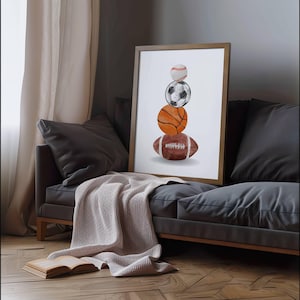 Pode incluir: Uma impress&atilde;o emoldurada com uma ilustra&ccedil;&atilde;o em aquarela de bolas esportivas empilhadas: beisebol, futebol, basquete e futebol americano. A obra de arte &eacute; exibida em um sof&aacute; cinza com um cobertor bege e um livro aberto.