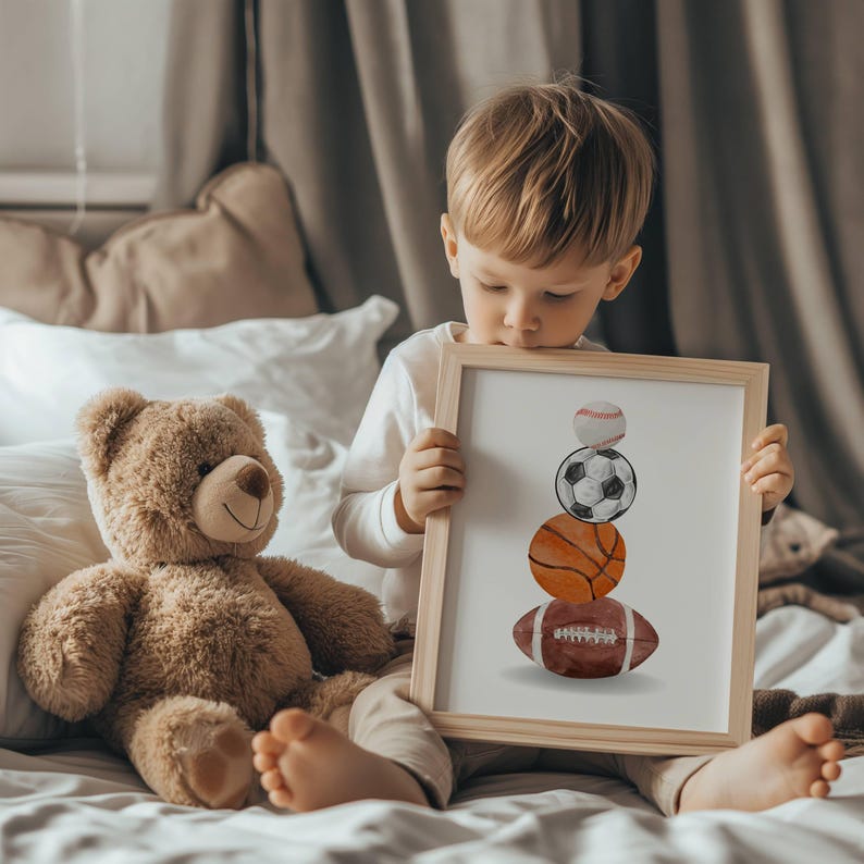 Pode incluir: Uma impress&atilde;o emoldurada com uma pilha de bolas esportivas em aquarela: beisebol, futebol, basquete e futebol americano. A impress&atilde;o tem uma moldura de madeira clara e &eacute; segurada por uma crian&ccedil;a. Um ursinho de pel&uacute;cia est&aacute; por perto.