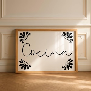 Puede incluir: Cartel enmarcado en madera con la palabra "Cocina" en escritura cursiva. El cartel tiene un fondo color crema con diseños florales negros en las esquinas. El marco es de color marrón claro y el cartel se muestra contra una pared blanca.