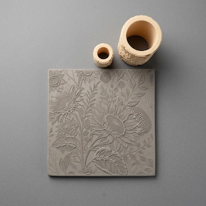 May include: A square gray clay tile featuring a detailed embossed sunflower and foliage pattern, accompanied by two cylindrical clay rollers of different sizes, each displaying a matching intricate floral relief design on its exterior surface.