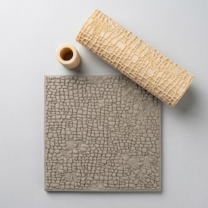 Könnte beinhalten: Eine beige, zylindrische Ton-Texturwalze mit Gittermuster, ein kleines passendes zylindrisches Werkzeug und eine quadratische, graue Tonplatte mit einem geprägten, unregelmäßigen, mosaikartigen Gittermuster, das dem Design der Walze entspricht.