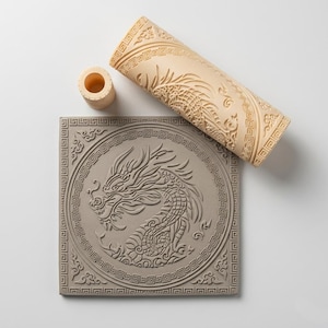 May include: A beige cylindrical patterned rolling pin and a small matching cylindrical connector piece positioned above a square grey clay tile featuring an embossed dragon design surrounded by a geometric Greek key border pattern.