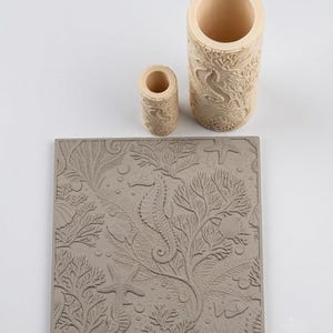 Puede incluir: Una placa cuadrada de arcilla gris y dos rodillos cilíndricos, uno grande y otro pequeño, todos con un patrón en relieve de caballitos de mar, coral, estrellas de mar y burbujas. Los rodillos son beige claro y la placa es gris.