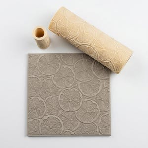 May include: A beige cylindrical rolling pin with a textured citrus slice pattern, a small matching cylindrical tool, and a square gray mat featuring an embossed pattern of overlapping citrus slices across the entire surface.