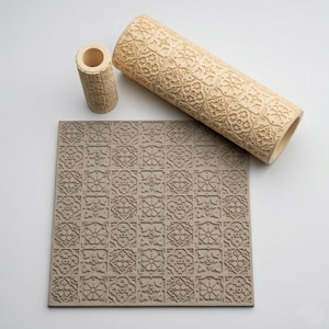 Puede incluir: Un rodillo cilíndrico de color beige y una herramienta cilíndrica más pequeña a juego, ambos con un patrón floral en relieve repetido, colocados sobre una lámina cuadrada plana con el mismo diseño floral en relieve en color gris claro.
