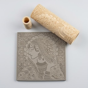 Puede incluir: Un rodillo cilíndrico de color beige claro con un intrincado patrón floral y de cachemira, una pequeña herramienta cilíndrica a juego y una baldosa de arcilla cuadrada gris con un perfil en relieve detallado de una persona con joyas.