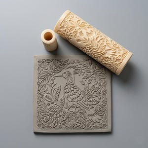 May include: A light-coloured cylindrical clay roller with an intricate floral and bird pattern, a smaller matching cylindrical tool, and a square grey clay tile embossed with the same detailed bird and foliage design in a relief texture.