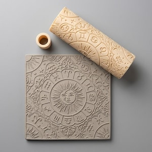 May include: A beige cylindrical rolling pin and a square flat tile, both featuring embossed zodiac signs and a central sun and moon face design. A small beige ring-shaped component is positioned near the top of the square tile.