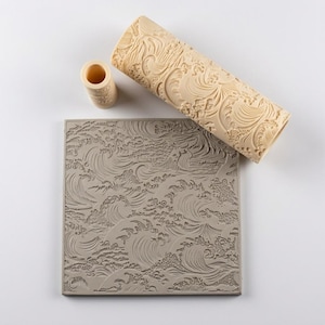 May include: A beige cylindrical patterned roller and a small matching cylindrical tool positioned above a square gray slab. The roller and slab feature a detailed, embossed design of stylized ocean waves with swirling water and foam textures.