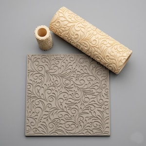 May include: A square gray clay texture sheet featuring an intricate embossed swirling leaf pattern, accompanied by a large cylindrical cream-colored rolling pin and a smaller matching cylindrical roller, both displaying the same embossed leaf design.